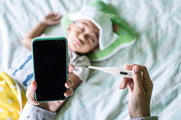 Mother holding a digital thermometer and smartphone to checking for her son sickness, illness, and fever flu while baby is slepping on a bed with wet cloth on his forehead.