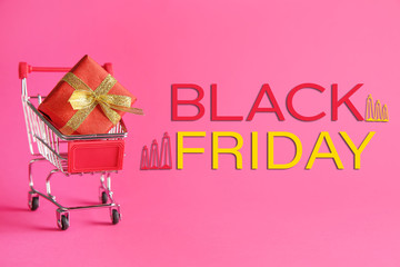 Small shopping cart with gift box and inscription BLACK FRIDAY on color background