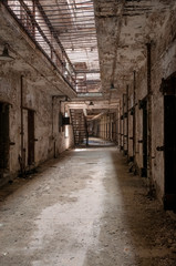 Hallway of an old abandoned prison showing doorways to cells, metal stairs, and bars on the ceiling.