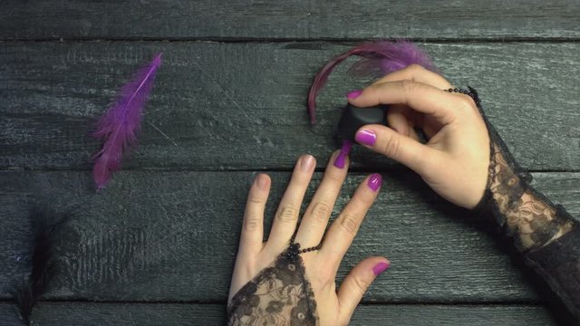 Nail design.  Girl paints her nails with purple gel polish on black wooden background among purple feathers. Graceful hands with a beautiful manicure on nails. 