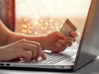 Cristmas online shopping soncept. Close-up woman's hands holding credit card and inputting card information using laptop keyboard for online shopping with festive lighting chain bokeh.