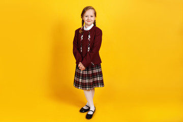 A full-length schoolgirl stands and looks at the camera. Cute little girl in uniform on a yellow background. - obrazy, fototapety, plakaty