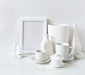 Fototapeta na wymiar Still life of white objects: cups with saucers, vases, an empty frame and candles on a white background. Place for text, soft light, high key.