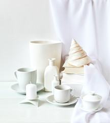 White still life with a stack of books, a sea shell, porcelain dishes and a candle against a background of white wall with a veil. Square image, place for text, high key, soft light