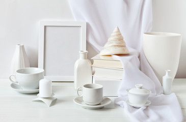 Still life in the style of a high key with a set of white objects: an empty frame, a stack of books, a seashell, porcelain dishes, vases of various shapes and a candle against a white wall with a veil