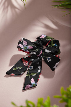 Object photo of a with black flower ornament scrunchie, decorating with big bow. The scrunchie is lying on a beige background. There are leaves in corners of a photo. 