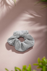 Object photo of a grey textured scrunchie. The scrunchie is lying on a beige background. There are leaves in corners of a photo. The scrunchie and leaves casting a shadow.