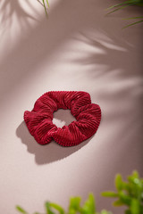 Object photo of a burgundy textured scrunchie. The scrunchie is lying on a beige background. There are leaves in corners of a photo. The scrunchie and leaves casting a shadow.