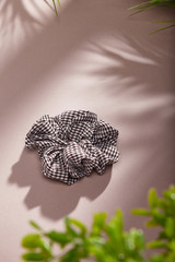 Object photo of a grey checked scrunchie. The scrunchie is lying on a beige background. There are leaves in corners of a photo. The scrunchie and leaves casting a shadow.