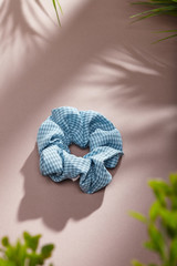 Object photo of a light blue checked scrunchie. The scrunchie is lying on a beige background. There are leaves in corners of a photo. The scrunchie and leaves casting a shadow.