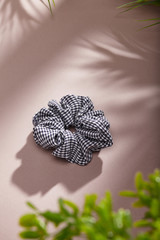 Object photo of a black checked scrunchie. The scrunchie is lying on a beige background. There are leaves in corners of a photo. The scrunchie and leaves casting a shadow.