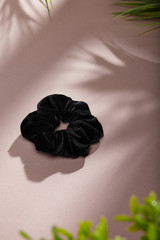 Object photo of a black velvet scrunchie. The scrunchie is lying on a beige background. There are leaves in corners of a photo. The scrunchie and leaves casting a shadow.