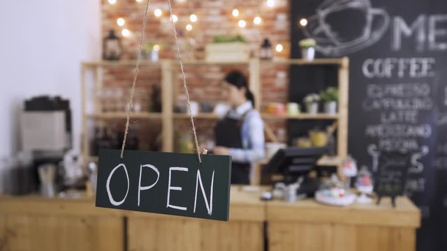 Open sign board close up in coffee shop door. bokeh view of asian korean woman cafe owner in wooden bar counter working. lady waitress in apron preparing in store in early morning coffeehouse.
