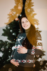 Pregnant woman posing near Christmas tree at home. Merry Christmas and Happy Holidays! Pregnancy, holidays, people and expectation concept.