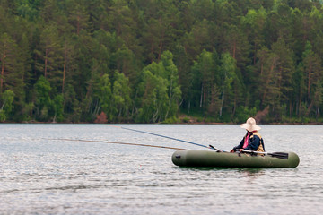 Fototapeta na wymiar A fisherman is fishing on fishing rods from a rubber boat with oars. The fisherman has a big sun hat. Two fishing rods. Selective focus. Trees on a blurred background.