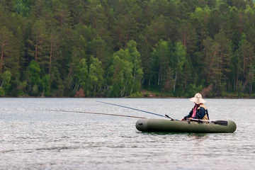 Foto op Canvas A fisherman is fishing on fishing rods from a rubber boat with oars. The fisherman has a big sun hat. Two fishing rods. Selective focus. Trees on a blurred background. © Vladimir Kazakov