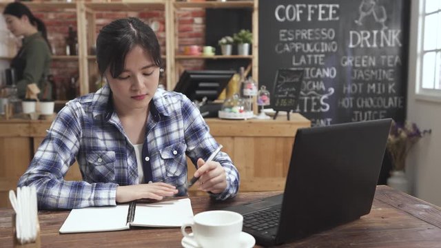 Young japanese woman sitting at table in cafe in front of laptop computer and making notes in notebook. girl student studying online website listening to tutor. education e learning concept