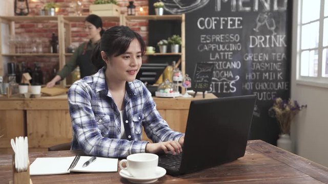 Female typing using laptop keyboard in cafe bar chatting with online friend. happy asian japanese woman smiling on notebook pc in coffee shop while busy waitress walking. internet friendship concept