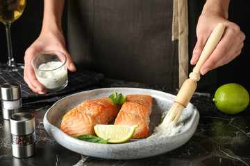 Woman adding sauce to cooked red fish on dark marble table, closeup