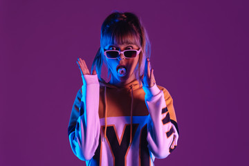 Amazed shocked pretty young fashion teen girl model wear glasses blowing bubble gum looking at camera stand at purple studio background, igen teenager in trendy stylish neon light concept, portrait