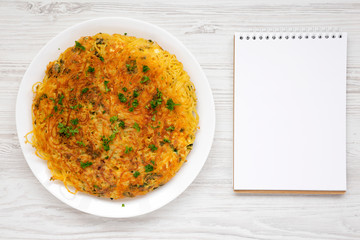 Homemade spaghetti omelette on a white plate, blank notepad over white wooden background, overhead view. Flat lay, top view, from above.