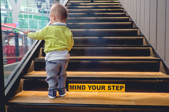 Cute Little Asian 18 Months / 1 Year Old Toddler Baby Boy Child Climbs Up The Wooden Stairs With The Mind Your Step Sticker Sign. Kid Trying To Walk Up Staircase - Selective Focus Y  By Yaoinlove