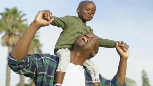 Young african boy sitting on his father's shoulders, holding hands smiling happy family quality time with blue sky