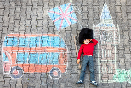 Happy little kid boy in british queen's guard soldier uniform having fun with London picture drawing with colorful chalks. With Big Ben, Union Jack and red bus as british doodles.