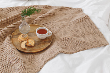 Breakfast in bed. Beige knitted blanket and wooden tray with cup of tea, cookies and vase with pine branche on bed with white linen. Cozy winter home decor.