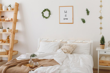 New year winter home interior decor. Holiday decorations. Stylish cozy white scandinavian bedroom: bed, knitted blanket, plaid, pillows, cushions, wreath, pine branches, little christmas tree in a pot