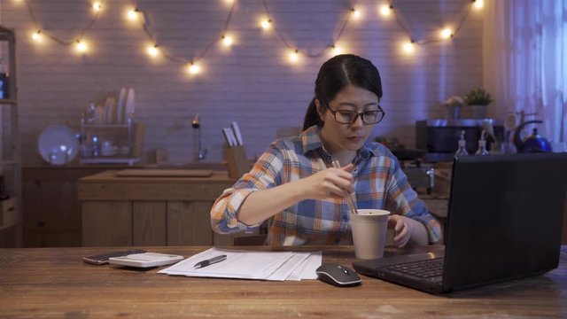 hungry asian korean hard working student study at home kitchen. college girl enjoy eating instant noodle in cup as late night supper by chopsticks. woman doing math counting on calculator and laptop