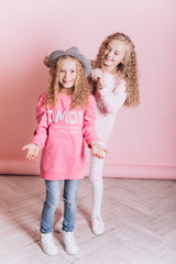 Close up portrait of young cute adorable caucasian sisters with curly hairstyle on soft pink background indoors in studio. Positive leisure concept, childhood happiness concept.