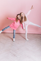 Close up portrait of young cute adorable caucasian sisters with curly hairstyle on soft pink background indoors in studio. Positive leisure concept, childhood happiness concept.