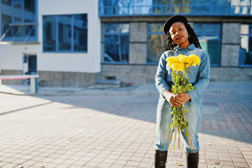 Fototapeta premium Stylish fashionable african american women in jeans wear and black beret with yellow flowers bouquet posed outdoor in sunny day against blue modern building.