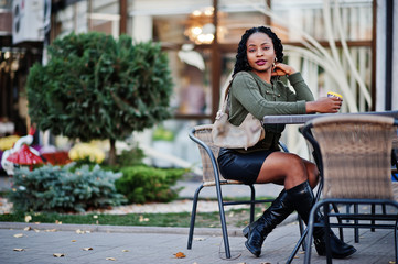 Stylish fashionable african american women in green sweater and black skirt posed outdoor cafe, sitting by table with cup of coffee.
