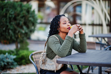 Stylish fashionable african american women in green sweater and black skirt posed outdoor cafe, sitting by table with cup of coffee.