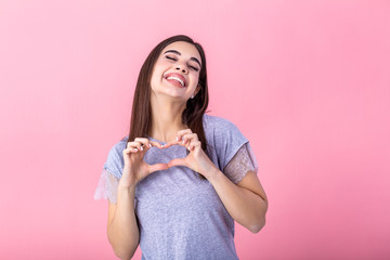 Portrait of a smiling young beautiful woman showing heart gesture with two hands and looking at camera isolated over pink background