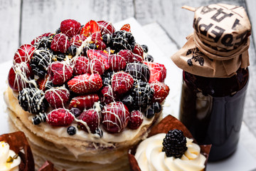 pancake cake with a cap of berries drenched in white chocolate on a wooden white table with a few Belgian muffins and a bottle of syrup