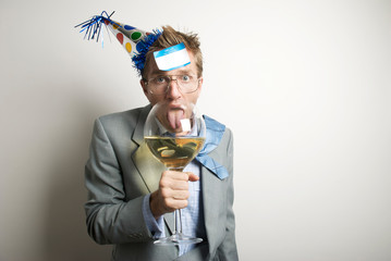 Drunk businessman wearing a New Year's Eve party hat with a name tag stuck to his forehead licking an oversized wine glass 