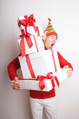 Holiday elf with big pointy ears and striped hat holding a towering stack of Christmas presents with a surprised expression