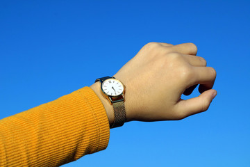 Girl dressed in yellow looking at the time on her gold and quartz watch. Blue sky background.