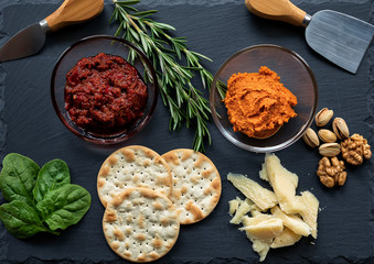 A dark cheese platter set with harissa and tomato sauce, parmigiano reggiano cheese, rosemary, spinach, walnuts and biscuits. Italian food. Flat lay. Traditional breakfast, snack or lunch.