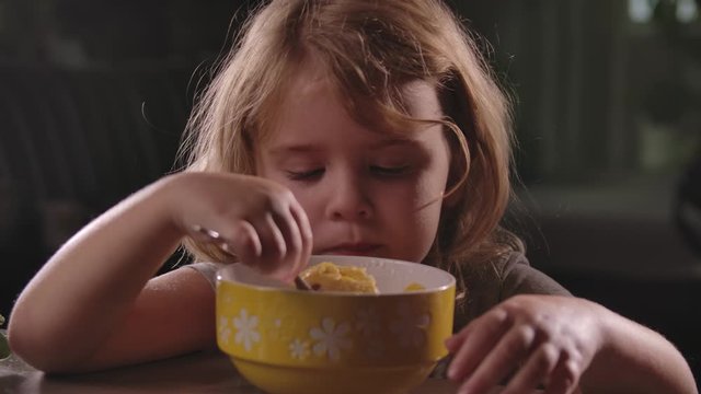 little cute girl with blond hair eats a spoon cereal with milk. Dry Breakfast with vitamins in the morning. The child eats at the table and laughs merrily. Portrait of a girl close-up.