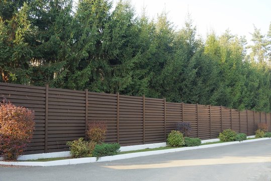 long brown wooden fence in front of a row of coniferous green trees by a gray road