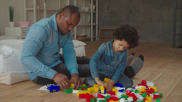 Engaged caring african dad and adorable preschool mixed race curly son building form colorful plastic blocks on floor at home. Multiethnic family playing developing game with constructor indoors.