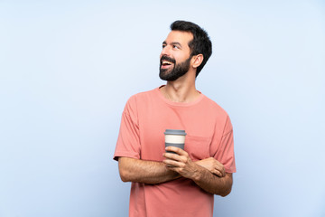Young man with beard holding a take away coffee over isolated blue background happy and smiling