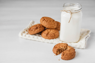 Milk in glass jar and oatmeal cookies near napkin on white table background. Copy, empty space for text