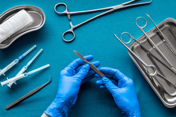 Preparing for plastic surgery. Doctor's hands takes scalpel on blue background with surgical tools top view - obrazy, fototapety, plakaty