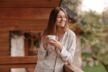 Fototapeta na wymiar Portrait of a happy smiling young woman in stylish nightwear with a cup of coffee or tea meeting the day standing on the hotel terrace with a gorgeous view of the mountains. place for inscription