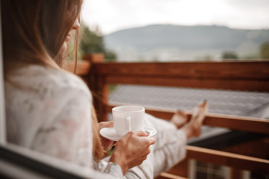 young woman in stylish nightwear enjoy drinking coffee or tea outdoor on balcony in the morning and looks at the mountains.
